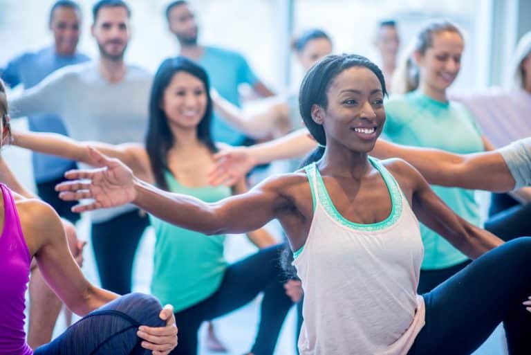doctor walking a patient through a balance test Focus on a woman in a floor class who is doing stretches with women and men. Everyone is balancing on one leg. The other leg is held up with one hand by the knee. The opposite arm on the standing leg is held outward and horizontally.